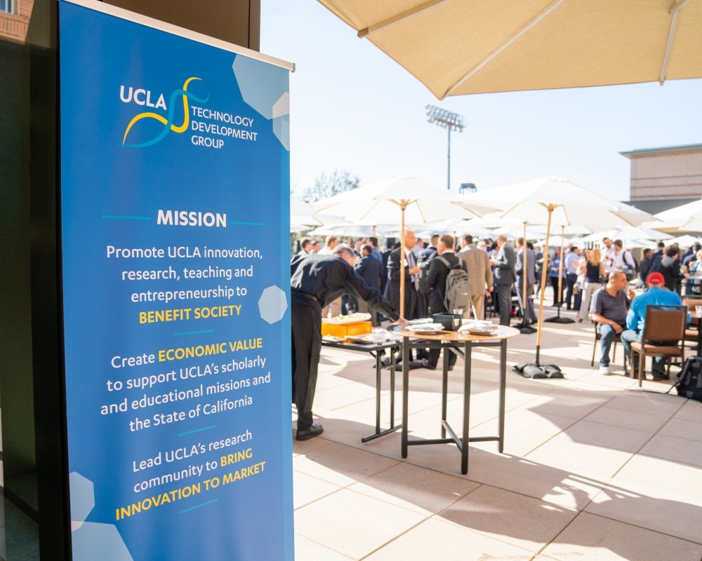 A crowd of attendees on an outdoor terrace under shade umbrellas, with a UCLA sign in the foreground