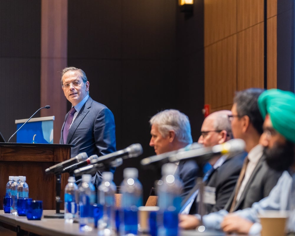 A speaker at the podium addressing the audience and a row of seated panel members